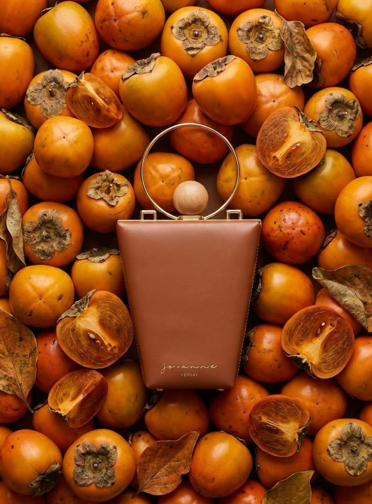 A structured brown leather handbag with a circular metal top handle and a wooden bead accent, centered on a dense background of whole and halved persimmon fruits