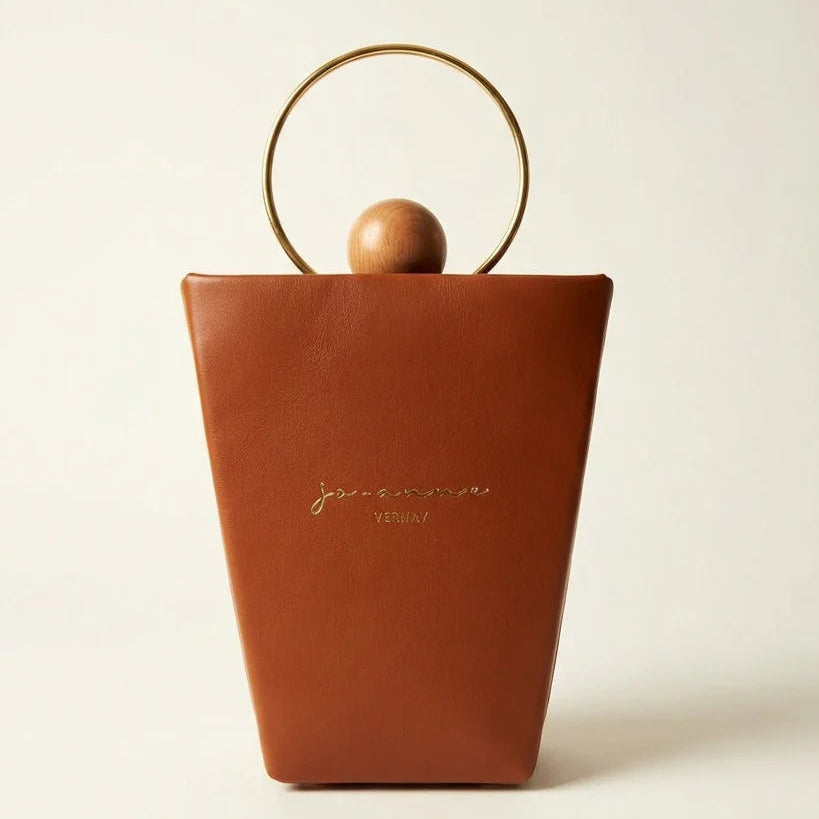 A structured brown leather handbag with a gold-tone circular metal handle and a small wooden sphere accent, featuring the brand name 'Jean Paul Gaultier' printed in gold on the front, set against a plain off-white background.