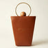 A structured brown leather handbag with a gold-tone circular metal handle and a small wooden sphere accent, featuring the brand name &