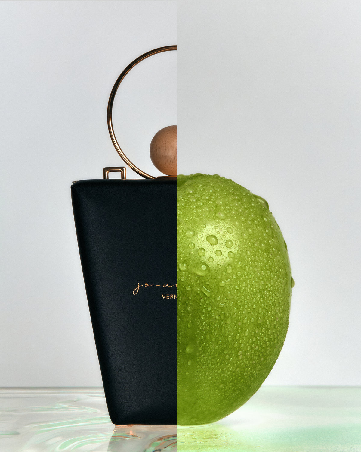 Black handbag with gold handle on a light gray background, split with a green fruit on a reflective surface.